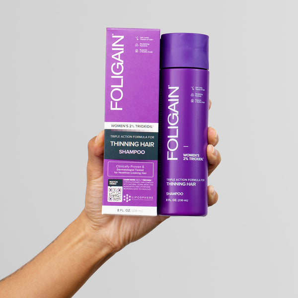 Foligain shampoo bottle and box held by a hand against a gray background
